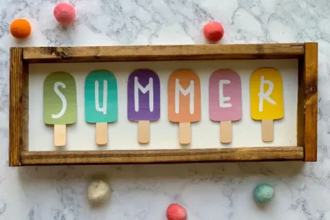 popsicles of different colors across a wooden shadow box. the popsicles spell summer.